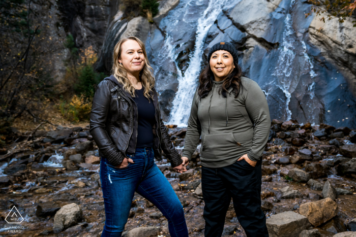 Couple Engagement Session at Colorado Springs Waterfall In Colorado Springs, an engaged couple stands side by side, holding hands in front of a cascading waterfall.