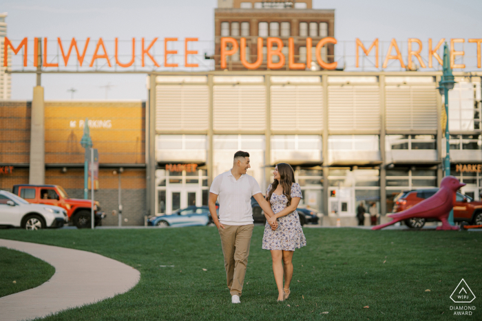 Milwaukee Couple's Sunset Stroll: Laughter, Love at Public Market In Milwaukee, Wisconsin, an engaged couple strolls together on the grass at sunset, exchanging jokes and laughter against the lively backdrop of the Milwaukee Public Market.
