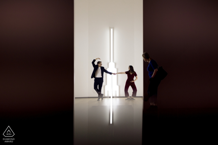 Engaged Couple's Graceful Ballet at National Gallery of Art At the National Gallery of Art in the City of Washington, an engaged couple gracefully dances, holding ballet poses as they hold hands. Framed by large doors and shot from another room, the scene exudes elegance and romance.