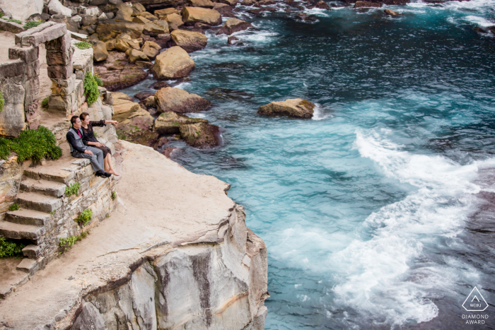 In Australia, an engaged couple sits on the steps of ancient ruins perched on the cliffs. Together, they watch the expansive blue sea below, with the powerful waves crashing against the rocks, creating a breathtaking and romantic scene.