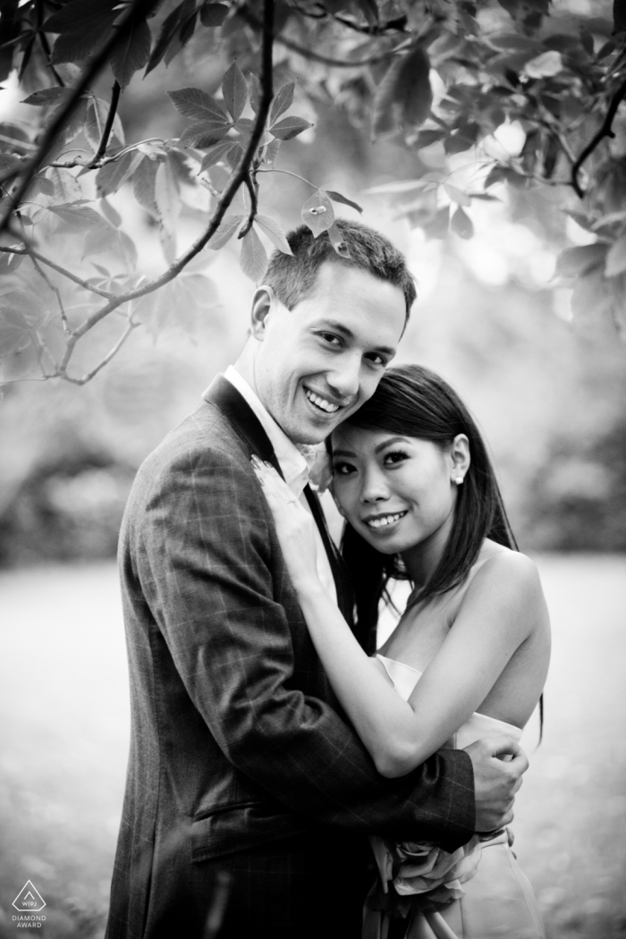 In London, UK, a couple is captured in a black-and-white portrait, arms lovingly wrapped around each other. Above them, muted foliage adds depth and layers to the image, in a serene and intimate atmosphere.