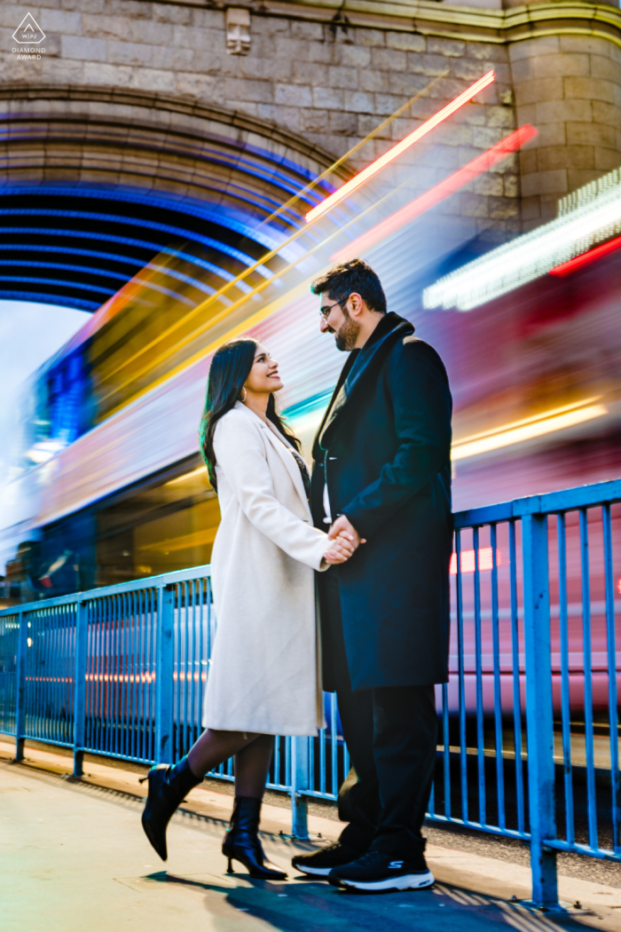 Engaged Couple at Tower Bridge, Standing Hand in Hand | Blurred Train Passing Adds Modern Energy and Sense of Movement to Portrait At the Tower Bridge, an engaged couple stands hand in hand. The dynamic backdrop of a blurred train passing beneath the iconic bridge adds a sense of movement and modern energy to their portrait.