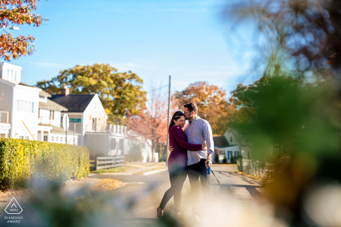 In Danbury, CT, an engaged couple stands on a sunlit residential street with their puppy. The warm sunshine highlights their joy and connection in their neighborhood.