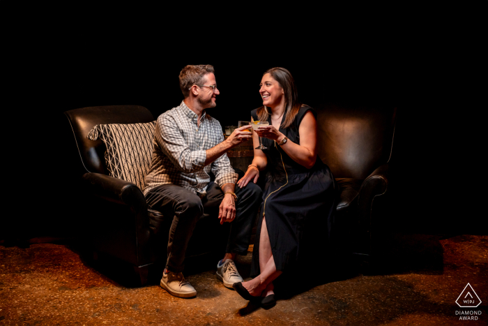 At SoNo 1420, a destination craft distillery in South Norwalk, CT, an engaged couple enjoys a cozy portrait session on a couch in a dimly lit area. Sharing a beverage, they bask in the warm and intimate atmosphere of the distillery.
