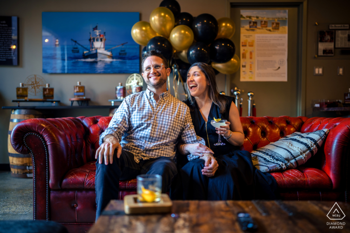 At SoNo 1420 America's Maritime Distillery in historic South Norwalk, CT, an engaged couple relaxes on a couch, watching TV and sharing laughter. The cozy setting within the craft distillery complements their joyful and intimate session together.