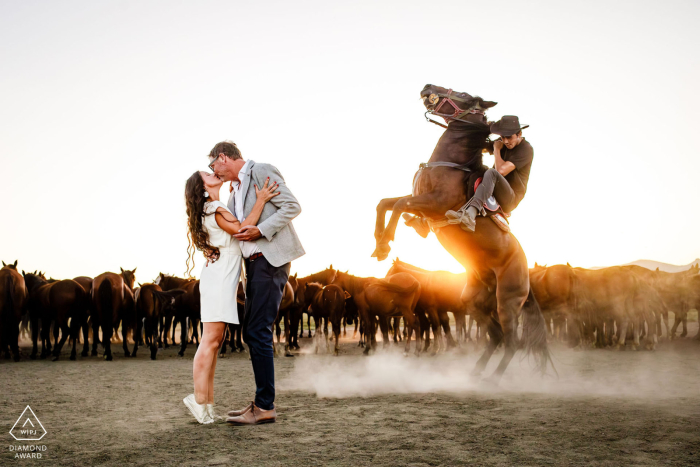 Engagement Session in Cappadocia, Kayseri, Turkey | Couple at Horse Ranch with Warm Glow from Low Sun | Cowboy on Rearing Horse Adds Dynamic Element In Kayseri, Turkey, during an engagement photo session in Cappadocia, a couple stands at a ranch with many horses. The low sun creates a warm glow, while to the right, a cowboy rides a horse rearing up, adding a dynamic element to the scene.