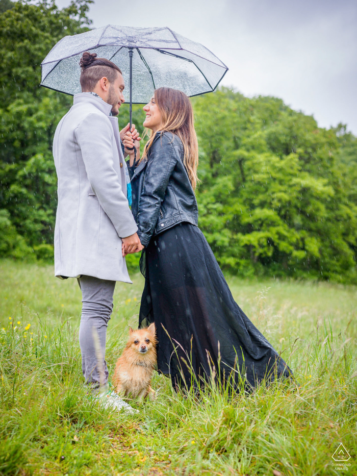 In Désert Jean-Jacques Rousseau, Seyssinet-Pariset, France, a couple stands on the grass under a single umbrella, with their dog sitting at their feet, creating a cozy and heartwarming scene.