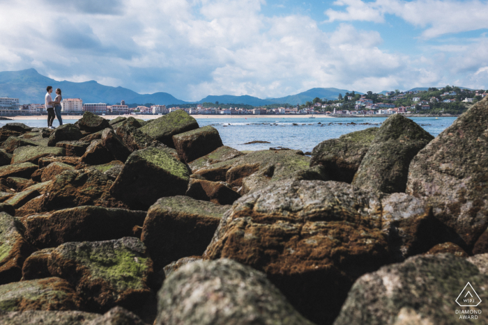 SAINT-JEAN-DE-LUZ, France — A couple stands on the rocks overlooking the bay of Saint-Jean-de-Luz, with the city and the La Rhune mountain in the distance for their engagement portrait.