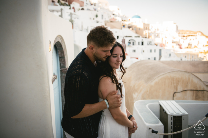 Santorini Embrace: An Affectionate Portrait of a Couple in Greece SANTORINI, Greece — A couple embraces above a village, with the groom-to-be hugging his fiancée from behind. This romantic engagement portrait captures their affection in Greece.