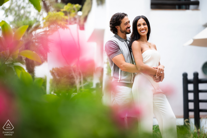 A couple poses at the Marbella Club, with him embracing her from behind. Both wear big smiles, surrounded by lush green foliage during their engagement session.
