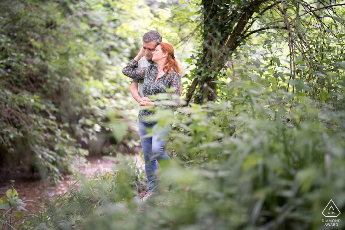 The engaged couple stands embracing near a tree and a small stream in a large park in Blagnac. They share an affectionate pose, surrounded by natural greenery during their engagement portrait session.