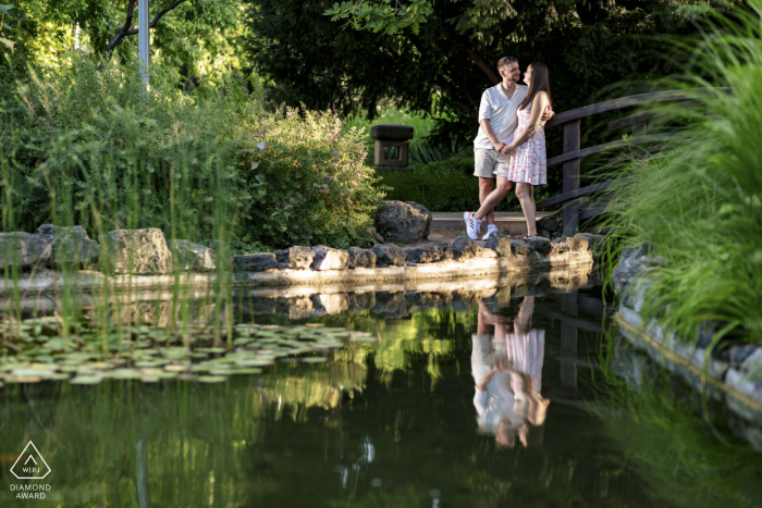 In Margitsziget, Hungary, a couple stands on a garden bridge, exchanging a loving gaze. Their reflection shimmers in the calm pond below, creating a serene and romantic engagement portrait.