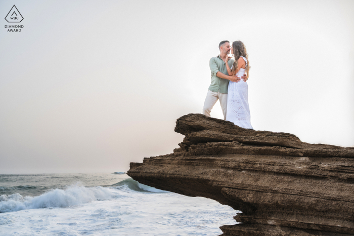 At Playa del Faro de Trafalgar, Cádiz, Spain, a pre-wedding portrait features the couple standing together on rocks overlooking the crashing waves of the sea.