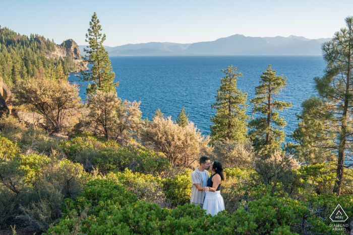 At South Lake Tahoe, CA, the engaged couple stands among the trees with a picturesque view of the sparkling lake visible through the foliage behind them.