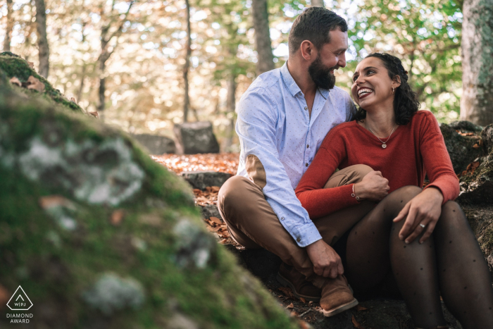 At Lac Chambon in France, the couple sits close together among moss-covered boulders in a lush forest, surrounded by the serene natural beauty.