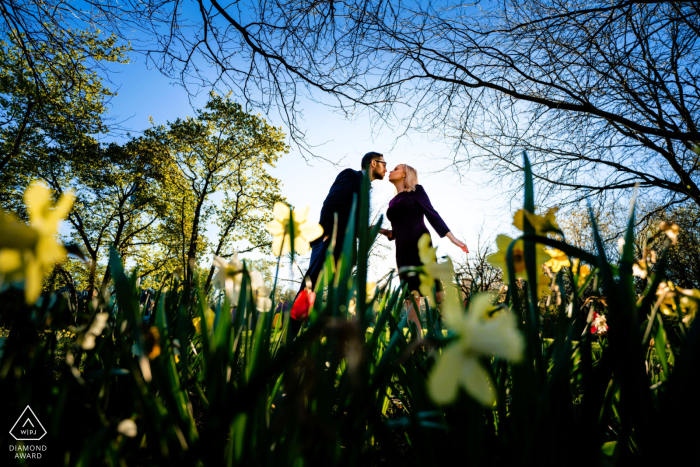 In Philadelphia, a couple leans in through tall grass and flowers, about to kiss, with trees and a blue sky above, captured in a portrait that highlights the photographer’s mastery of location lighting.