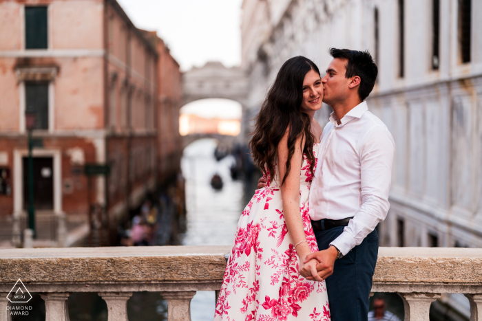An engaged couple hugs and kisses while standing on a footbridge over a canal in Venice, Italy, during a portrait session with a WPJA photographer.