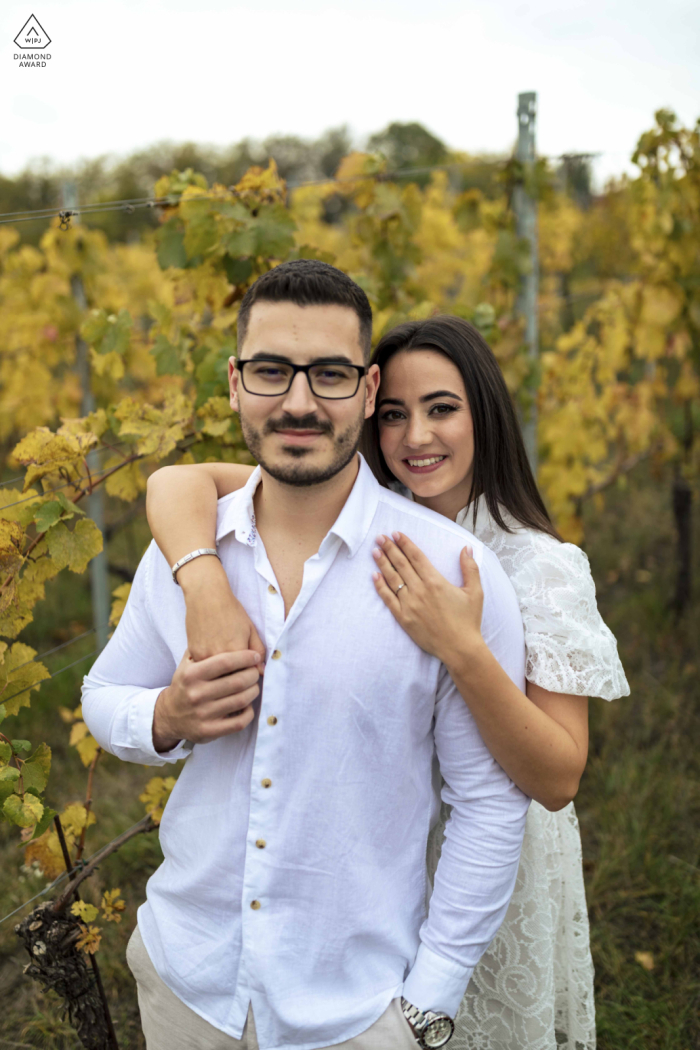 A smiling couple stands close together in an autumn vineyard in Etyek, Hungary; she embraces him from behind as both look at the camera, quietly celebrating their love and engagement.