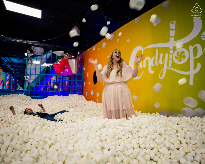 A couple celebrates joyfully in a marshmallow-themed ball pit in Manhattan, NY, tossing and surrounded by falling fluffy balls, creating a whimsical and playful engagement portrait.