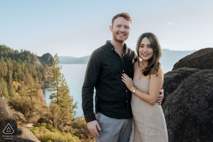 Engaged Couple Smiling Side By Side At South Lake Tahoe With Scenic Mountain And Lake View Behind Them The couple stands side by side at South Lake Tahoe, smiling at the camera with the scenic view stretching out behind them, captured in a joyful and scenic engagement portrait.