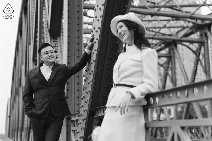 The groom to be leans against a bridge beam at the Bund in Shanghai, China, gazing at his future bride with adoration as she laughs, enjoying a beautiful and timeless afternoon together in the city.
