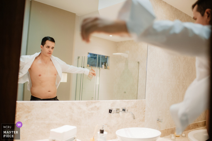 Photo by: <a href="https://www.wpja.com/contests/2018-v01-photography-competition/best-wedding-photographers/2167981">Gustavo Lucena - Brasilia Distrito Federal</a> Brazil groom looking at himself in the mirror as he puts a shirt on before the wedding