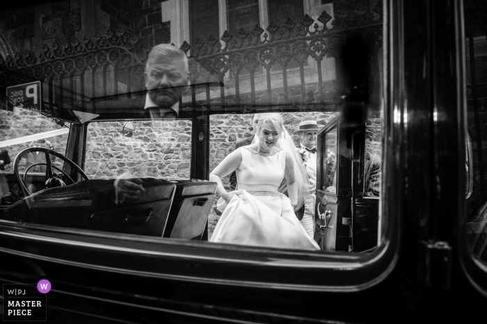 Photo by: <a href="https://www.wpja.com/contests/2018-v01-photography-competition/best-wedding-photographers/2173726">Nick Despres - Guernsey Guernsey</a> Guernsey bride getting into the vintage wedding car while holding her dress - black-and-white photography