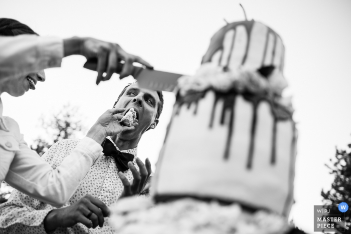 Foto di: <a href="https://www.wpja.com/contests/2018-v07-photography-competition/best-wedding-photographers/2228076"> Carl Bower - Denver Colorado </a> La sposa maneggia coltello e torta alle nozze all'aperto a Boulder, Colorado. La sposa impone la torta nuziale nella bocca dello sposo. Sunrise Amphitheatre, Boulder, Colorado