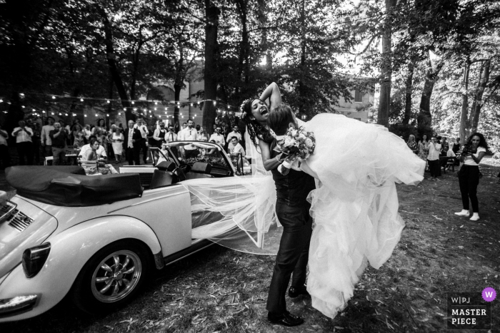 Foto von: <a href="https://www.wpja.com/contests/2018-v09/best-wedding-photographers/2238866"> Melissa Iannace - San Vito von Spilamberto Modena </a> Modena Bräutigam hebt die Braut in die Luft draußen am Hochzeitsempfang beim Verlassen des konvertierbaren VW-Käfers.
