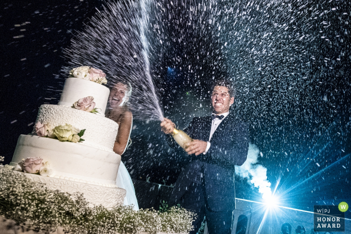 Foto door: <a href="https://www.wpja.com/contests/2018-v09-photography-competition/best-wedding-photographers/2239801"> Luigi Rota - Comomeer Lecco </a> Groom knalt een fles champagne naast de taart - Lecco trouwfotograaf