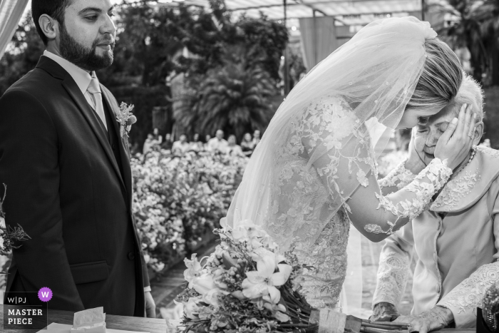 Photo: <a href="https://www.wpja.com/contests/2018-v10-photography-competition/best-wedding-photographers/2252456"> Clara Sampaio - Rio de Janeiro Rio de Janeiro </a> Photo noir et blanc du marié se tenant à côté de la mariée alors qu'elle embrasse une femme âgée devant un photographe de mariage à Rio de Janeiro, au Brésil.