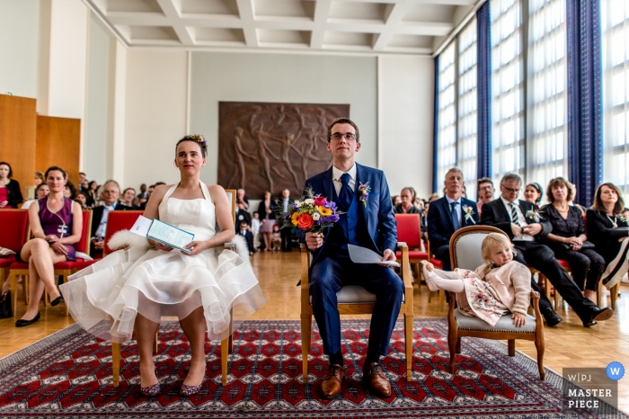 Brest, Frankreich Hochzeitszeremonie Foto von Braut, Bräutigam und Blumenmädchen sitzen in Stühlen mit Gästen hinter ihnen Hochzeitszeremoniefoto Brests, Frankreich der Braut, des Bräutigams und des Blumenmädchens, die in den Stühlen mit Gästen hinter ihnen sitzen.