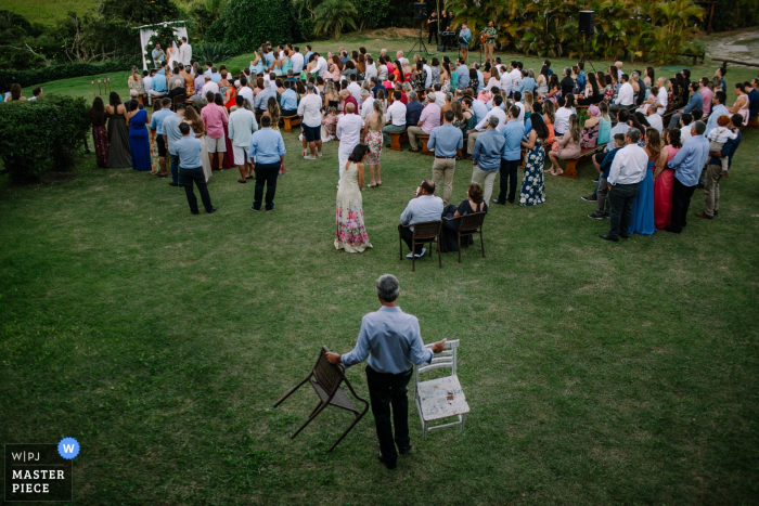 Praia do Rosa - Santa Catarina - Brasil  Wedding Photojournalist | a man carries extra chairs to the outdoor wedding ceremony on the grass lawn