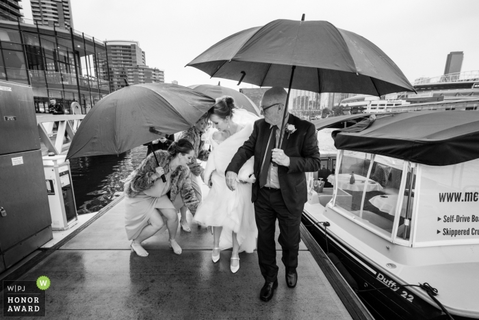 Foto: <a href="https://www.wpja.com/contests/2018-v12-Fotowettbewerb/Best-wedding-Photographers/2276091"> Alan Rogers - Doncaster East Victoria </a> Australien-Brautparty, die vom Boot im Regen mit Regenschirmen bei den Melbourne Docklands erhält.