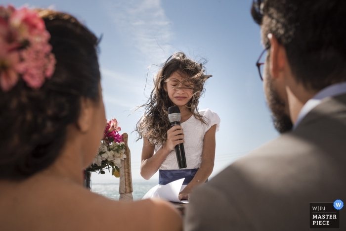 Búzios, Rio de Janeiro, Brasil Fotojornalista de casamento | uma menina chora enquanto segura um microfone enquanto fala com a noiva e o noivo durante esta cerimônia de casamento ao ar livre