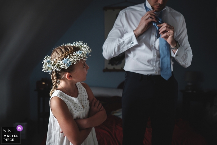 Photo by: <a href="https://www.wpja.com/contests/2018-v13/best-wedding-photographers/2283166">Didier Ropers - Lorient </a> Morbihan Wedding Photojournalist | flower girl in her dress with flowers in her hair watches a man tie his tie