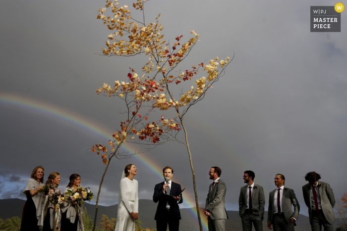 Ceremonia de bodas al aire libre de ripton en Vermont bajo dos árboles cruzados y un hermoso arco iris detrás de ellos