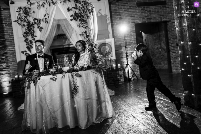 Photo by: <a href="https://www.wpja.com/contests/2018-v15/best-wedding-photographers/2307776">Wenjie Han - Long Island City New York</a> Black and white photo of a small boy with the camera creating an image of the bride and groom at their head table during the reception