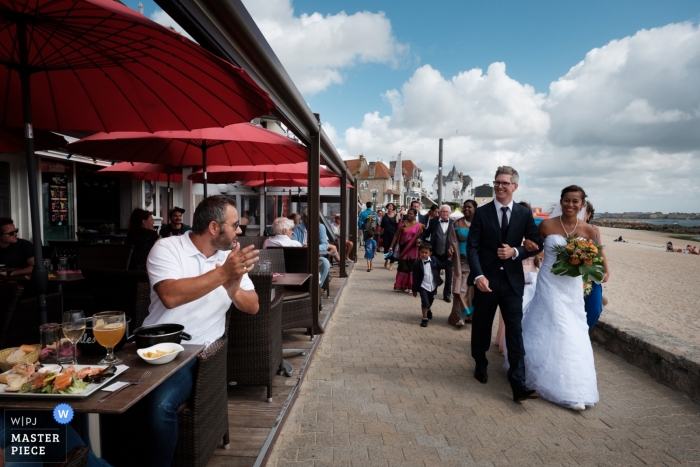 Photo by: <a href="https://www.wpja.com/contests/2018-v15/best-wedding-photographers/2315676">Didier Ropers - Lorient </a> Morbihan wedding photojournalism image of a couple walking past diners at an outdoor café in Brittany