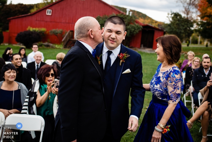 Documentaire huwelijksfoto van een bruidegom die een kus op de wang van zijn vader krijgt bij een openluchtceremonie in Charlotte, Vermont bij een rode schuur