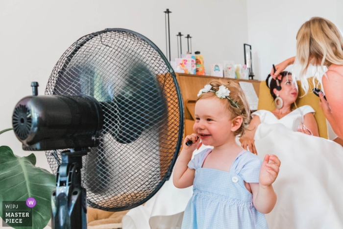 Photo by: <a href="https://www.wpja.com/contests/2018-v17/best-wedding-photographers/2341356">Natalja van Ommeren - Zwartewaal Zuid Holland</a> Wedding pictures of a young flower girl cooling off with the fan as the bride has make applied - Zuid Holland photographer