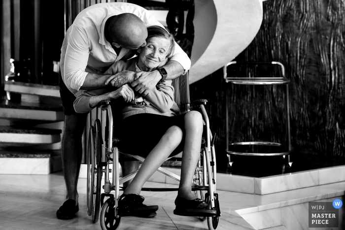 Photo by: <a href="https://www.wpja.com/contests/2018-v18/best-wedding-photographers/2360991">Robert David - București</a> Bucuresti wedding photograph of an elderly woman in a wheelchair receiving a hug and a kiss from a man behind her