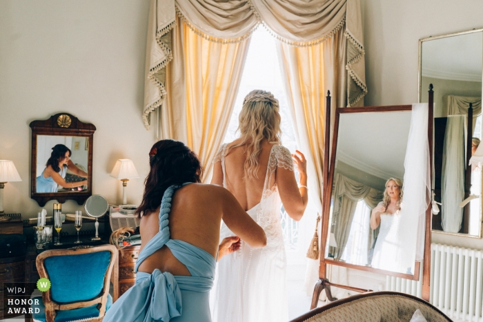 Zdjęcie: <a href="https://www.wpja.com/contests/2018-v20-photography-competition/best-wedding-photographers/2389971"> Hannah Walker - Merriott Somerset </a> Ślub przygotowuje się do sesji zdjęciowej w North Cadbury Court, Somerset UK - Druhna pomaga pannie młodej w sukni ślubnej z odbiciami w lustrach