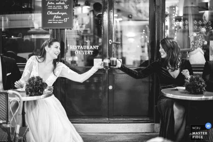 Hochzeitsfoto einer Braut, die mit ihrer Freundin im Café anstößt | Momente des Hochzeitstags, festgehalten in Paris, Frankreich