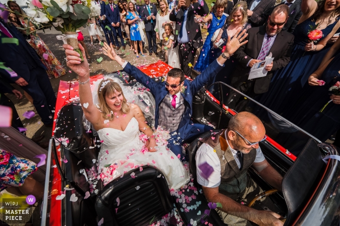 Fotografia di matrimonio degli sposi che si allontanano in auto decappottabile | Momenti del giorno del matrimonio catturati nel Northamptonshire, nel Regno Unito