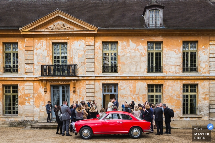 Photo by: <a href="https://www.wpja.com/contests/2019-v01/best-wedding-photographers/2425671">Ronan Jégaden - Athis-Mons </a> Paris Ceremony photograph of a red car, the bride and groom, and guests
