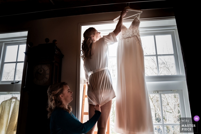 Foto: <a href="https://www.wpja.com/contests/2019-v01/best-wedding-photographers/2437866"> Thomas Jongbloed - Utrecht Utrecht </a> Spoorwegmuseum Utrecht, NL - Die Braut bereitet ihr Kleid vor, damit es fotografiert werden kann, während es im Raum hängt. Ihre Freundin kümmert sich um ihr kurzes Nachtkleid, damit es nicht krabbeln kann und zieht es herunter.