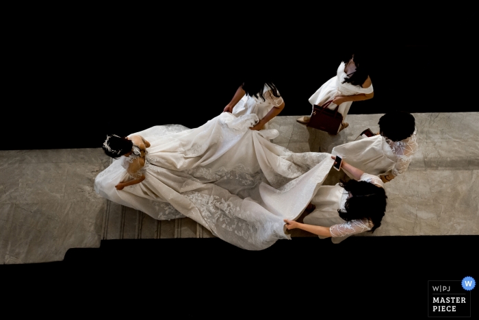 Photo by: <a href="https://www.wpja.com/contests/2019-v04/best-wedding-photographers/2465491">Qi Xiang - Nanping </a> Bridesmaids help the bride with her dress as she walks down the hotel stairs toward the ceremony in this photo by a Fujian, China wedding photographer.