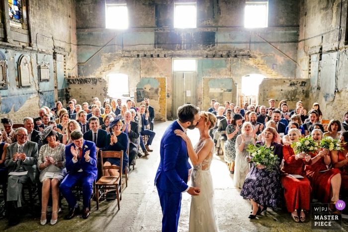 Photo by: <a href="https://www.wpja.com/contests/2019-v04/best-wedding-photographers/2467421">Andrew Billington - Leek Staffordshire</a> AMP Studios, Peckham Photo of the First Kiss with Family and Guests Seated.