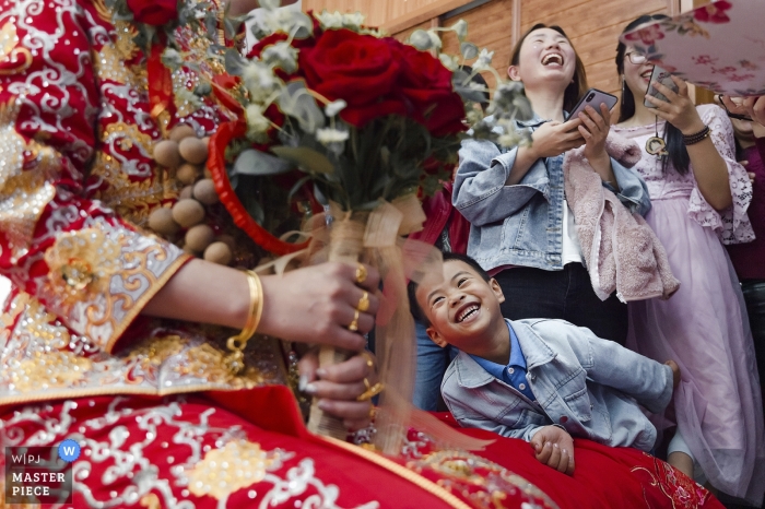 Photo by: <a href="https://www.wpja.com/contests/2019-v04/best-wedding-photographers/2468131">Cen Lin - Fuzhou </a> Photo of a young boy admiring the bride's colorful dress and bouquet at a Fuzhou home by a China wedding photographer.